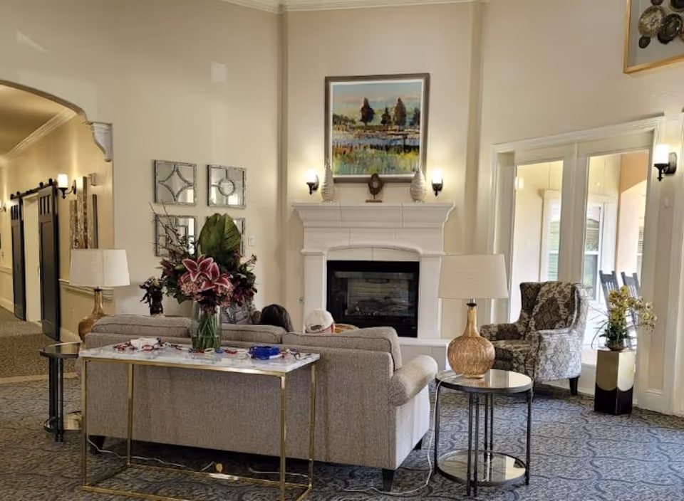 A cozy senior living facility common area with a beige sofa, a floral armchair, a fireplace with a painting above it, and side tables with lamps. There is a large flower arrangement on a console table behind the sofa. The room has carpeted floors and large windows letting in natural light.