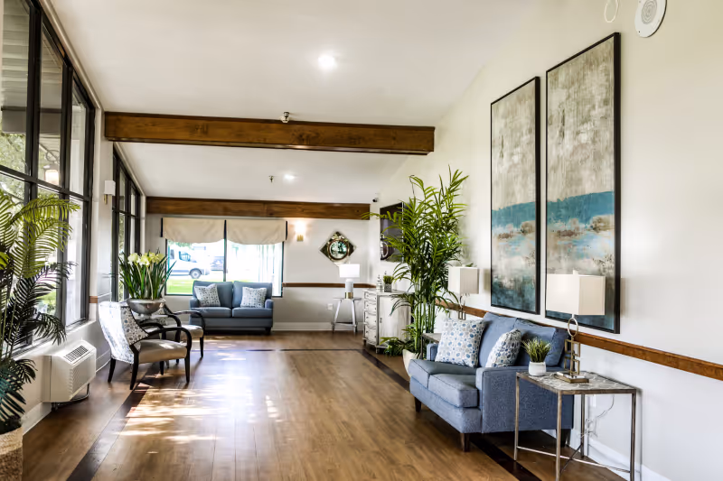 Bright, furnished lounge with sofas, armchairs, large windows, plants, and wall artwork.