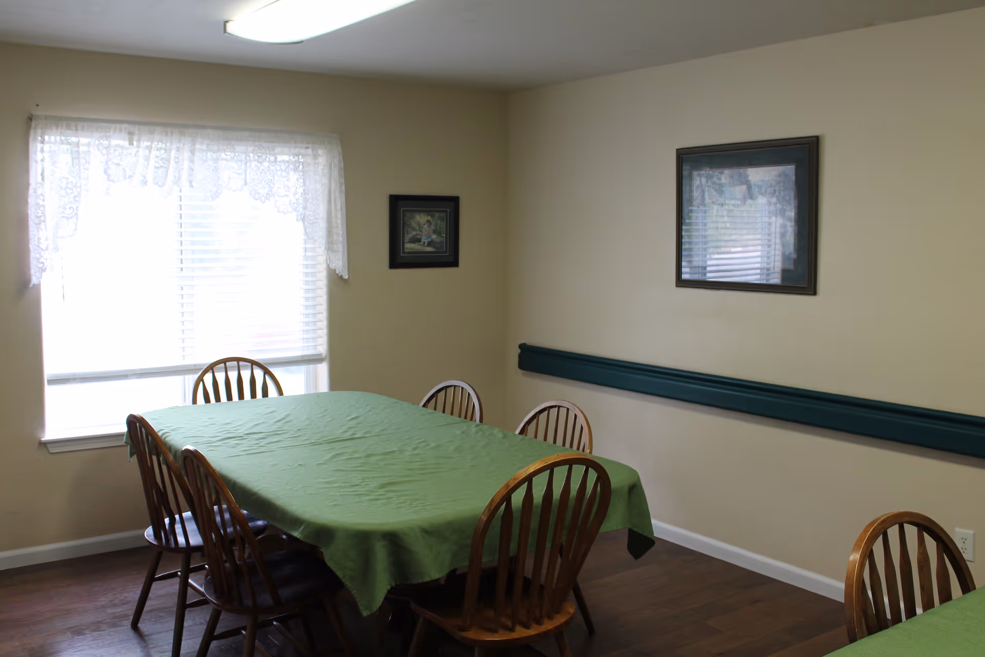 Small dining room with wooden chairs around a table covered by a green tablecloth, a window with lace curtains, and framed pictures on beige walls.