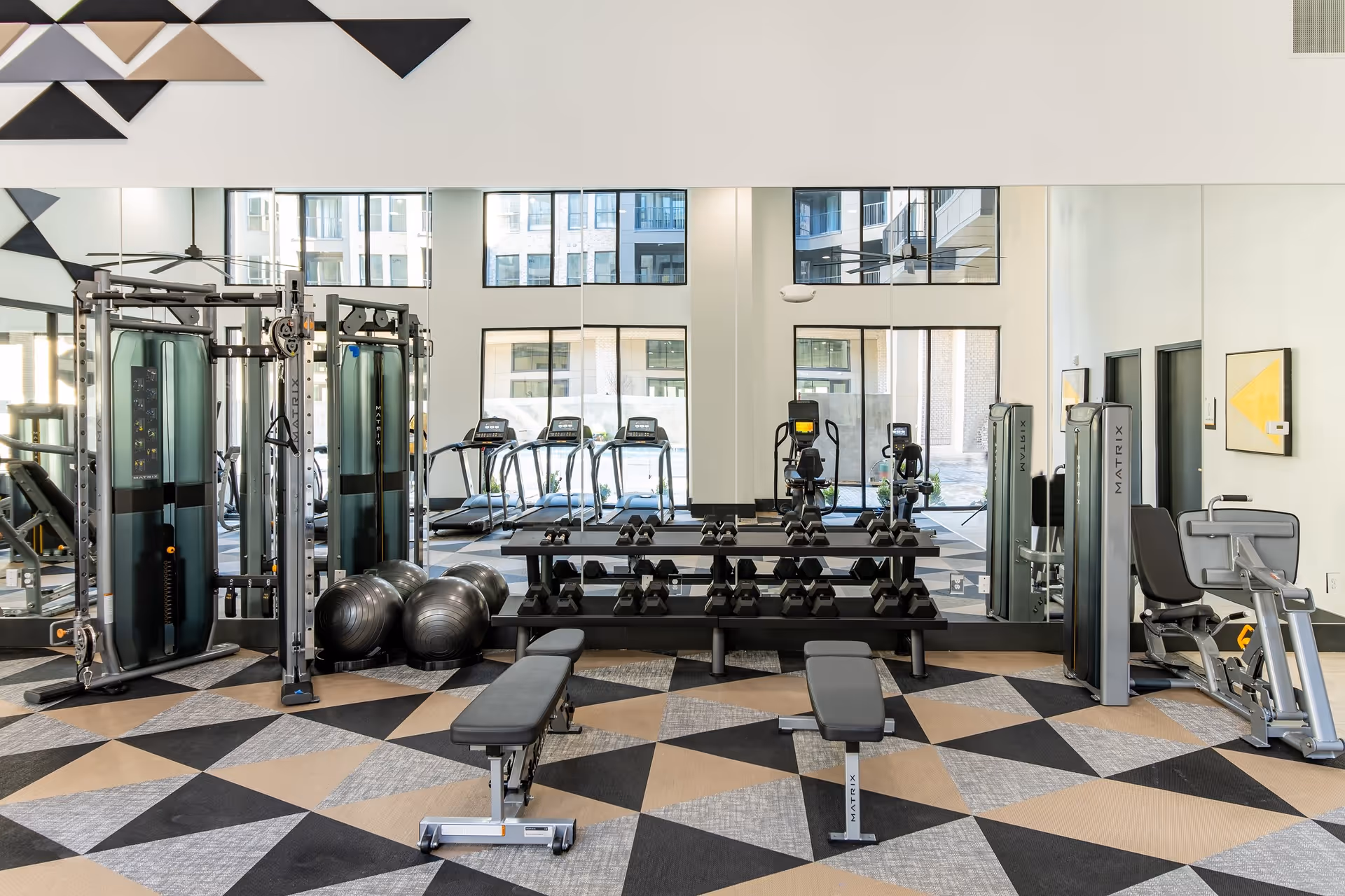 A modern fitness room with various exercise equipment including weight benches, dumbbells on racks, exercise balls, treadmills, and strength training machines. The room has large windows and mirrors on the walls, with a geometric patterned carpet in shades of black, beige, and gray.