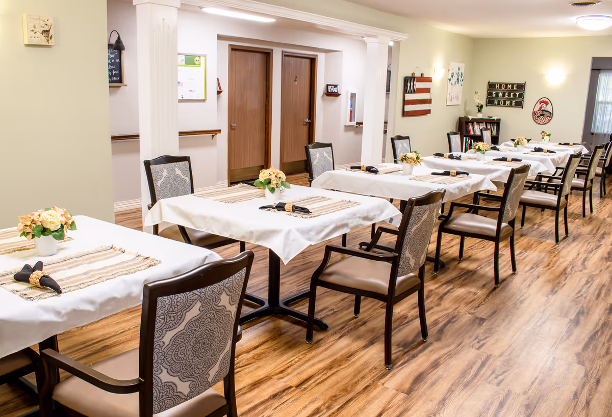 Dining room with rows of tables set with white tablecloths, floral centerpieces, and chairs on wood flooring.