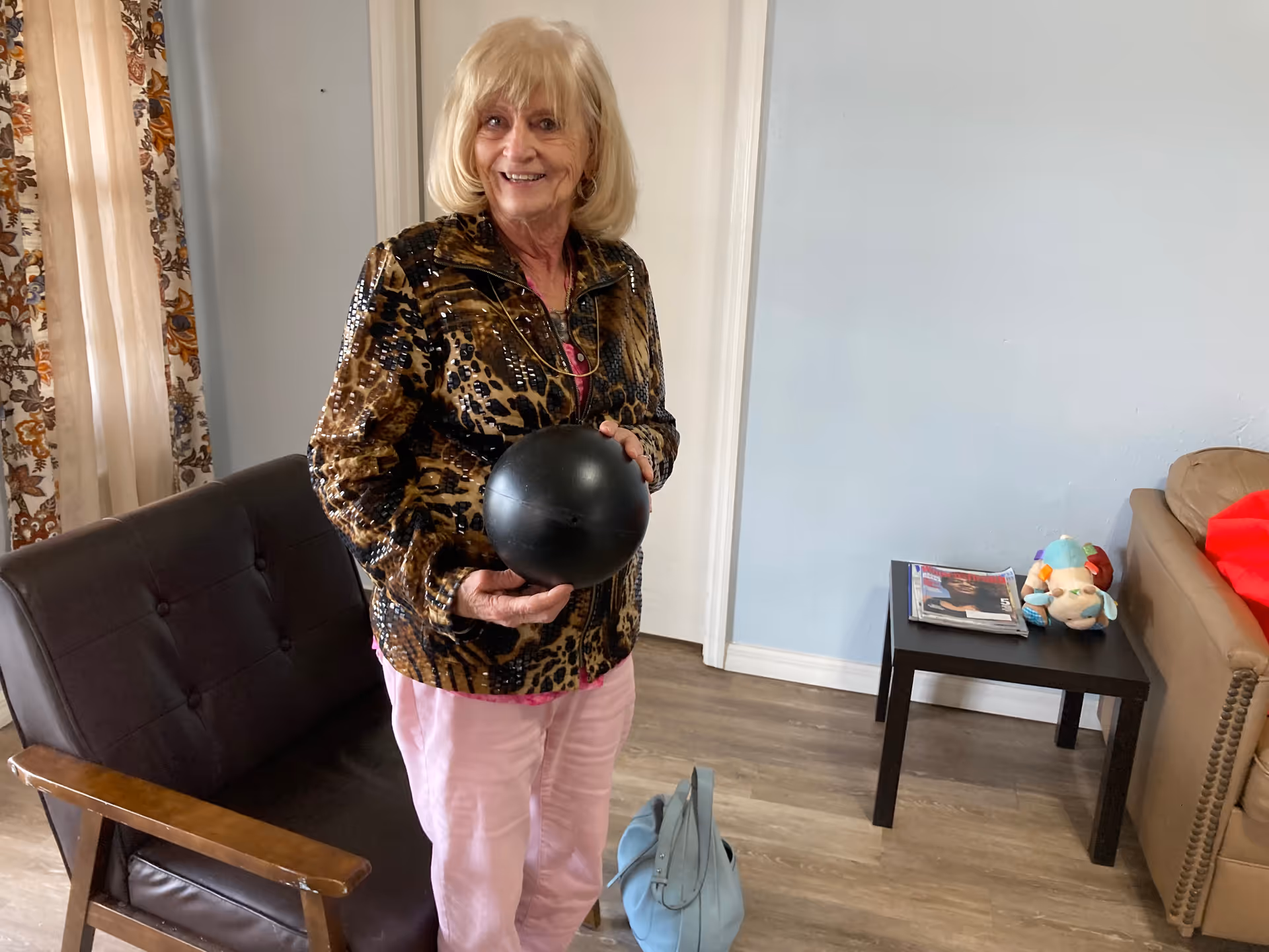 An elderly woman with blonde hair wearing a patterned jacket and pink pants is standing indoors holding a black ball. She is smiling and standing next to a brown cushioned chair with wooden armrests. Behind her is a small black side table with magazines and a stuffed toy on it, and a beige couch with a red blanket. The room has light blue walls and wood flooring.