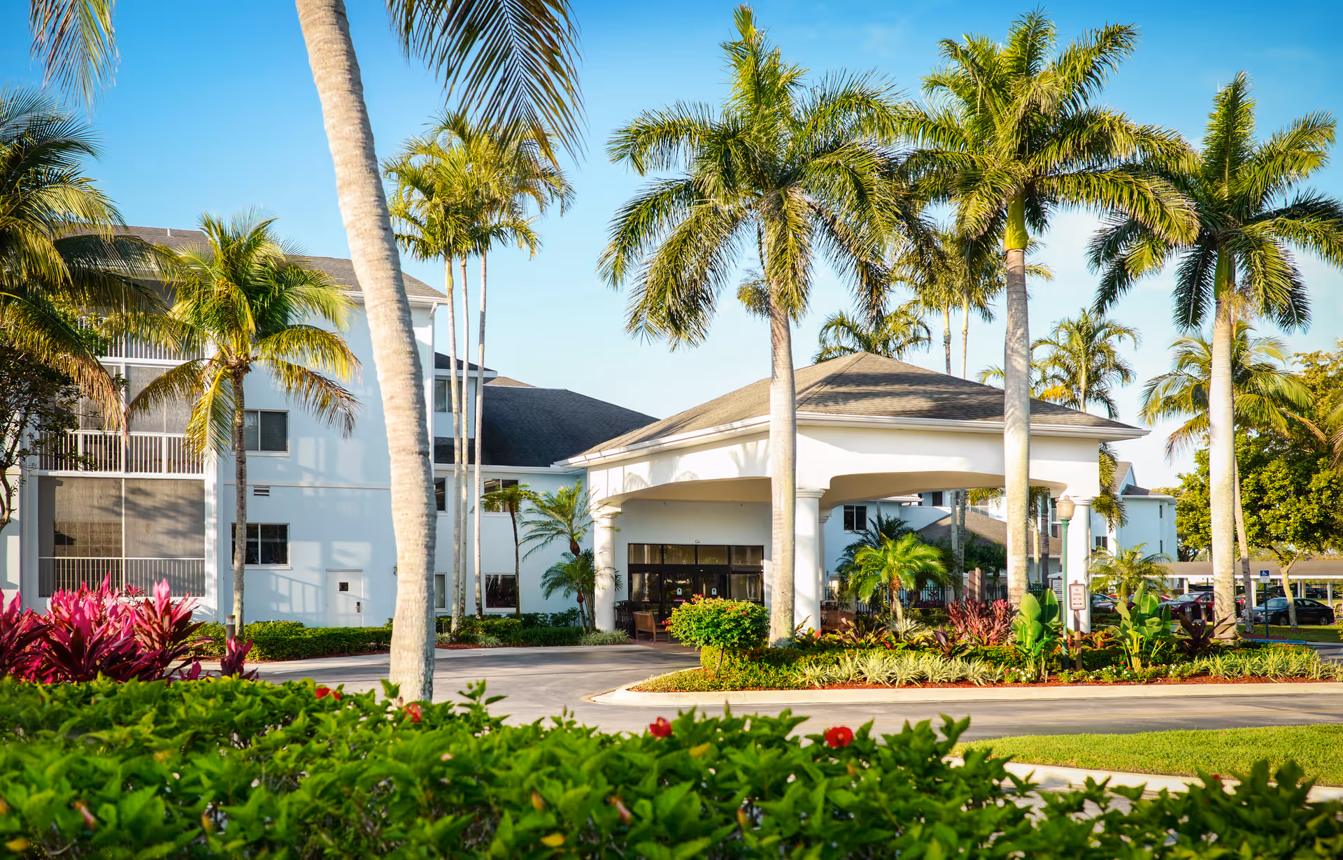 White senior living facility with palm trees, lush landscaping, and a covered entrance