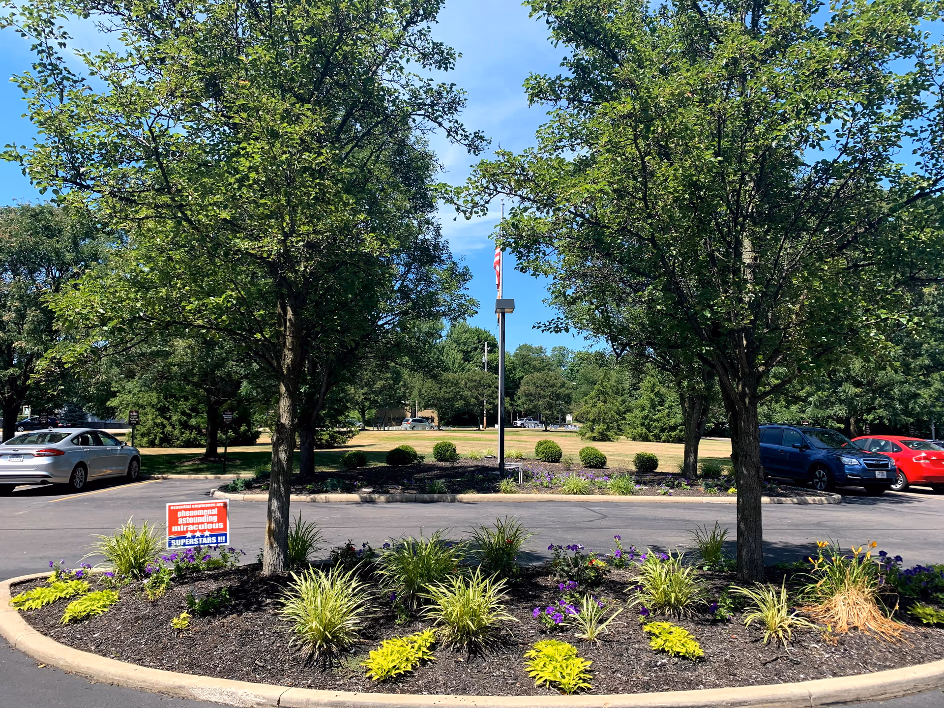 A landscaped roundabout with two trees and various green plants and flowers in the center, surrounded by a parking lot with several parked cars. In the background, there is a grassy area with more trees and an American flag on a pole under a clear blue sky.