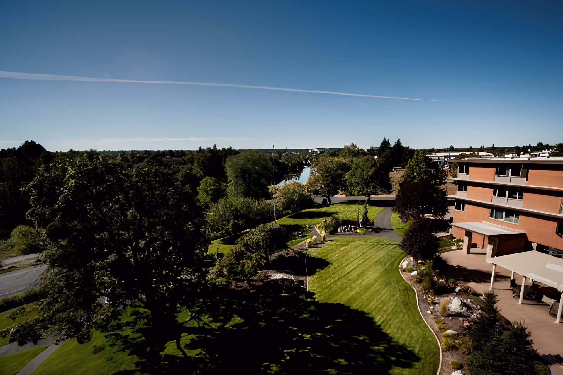 Aerial view of Riverview Lutheran Retirement Community of Spokane showing a large green lawn with trees, a winding pathway, and a multi-story brick building on the right side. The background features a river and dense forest under a clear blue sky.