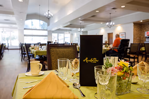 A dining room in a senior living facility with tables set for a meal. The table in the foreground has a yellow napkin, glassware, silverware, a coffee cup, a floral centerpiece, and a black menu with the Acclaim Living logo. In the background, there are more tables and chairs, large windows letting in natural light, and a few people seated.