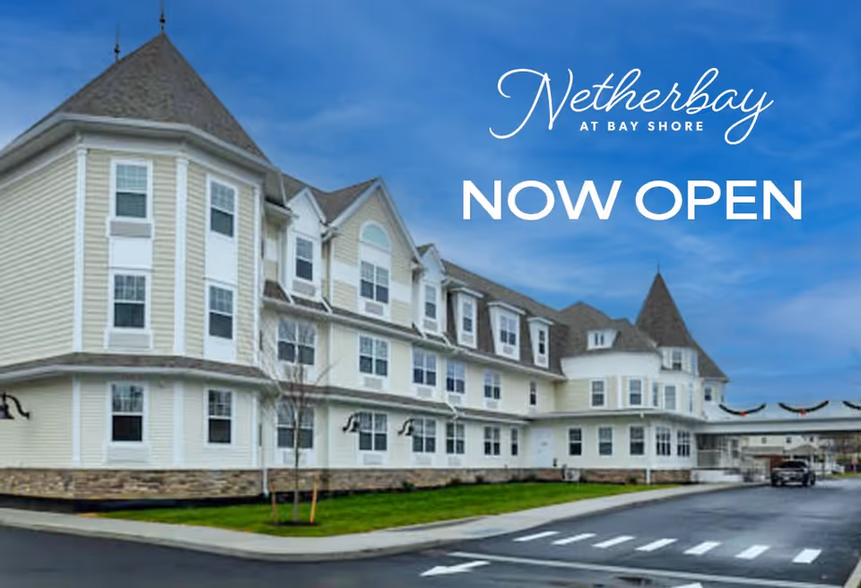 Exterior view of a large, multi-story senior living facility building with beige siding, white trim, and multiple windows under a blue sky. The building has a distinctive turret and a covered entrance with a driveway. Text overlay reads 'Netherbay at Bay Shore NOW OPEN'.