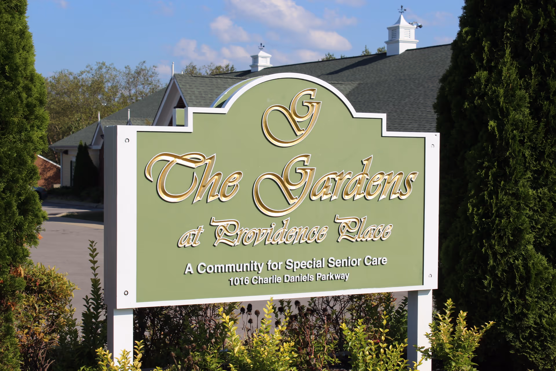 A green and white sign for The Gardens at Providence Place, a community for special senior care located at 1016 Charlie Daniels Parkway, surrounded by greenery and trees with a building and blue sky in the background.