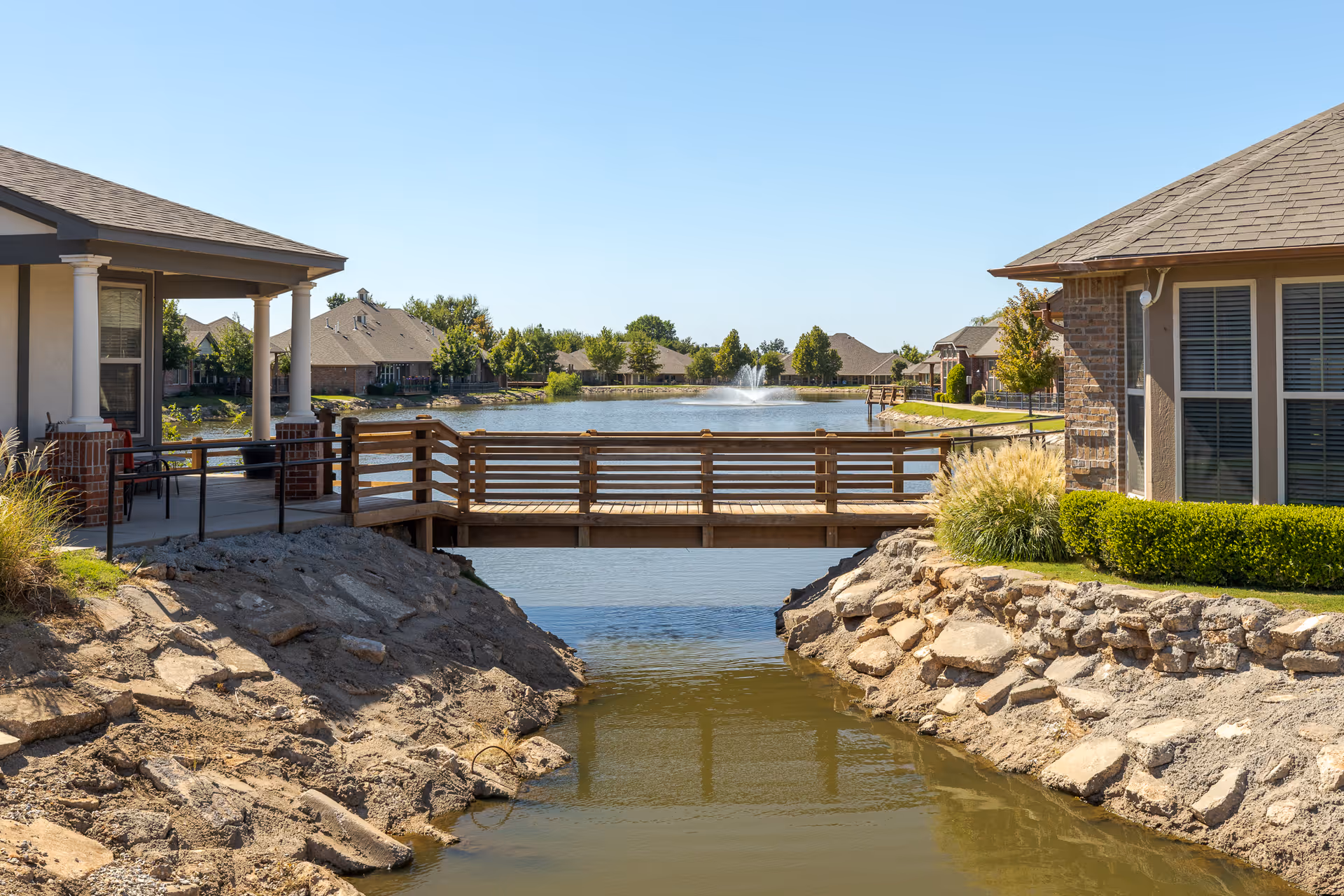 A wooden footbridge over a narrow water channel leading to a larger pond with a water fountain in the center, surrounded by residential buildings and greenery under a clear blue sky.