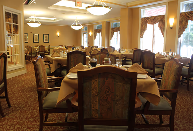 A well-lit dining room with multiple tables set with tablecloths, napkins, glassware and upholstered chairs beneath ceiling lights and large windows.