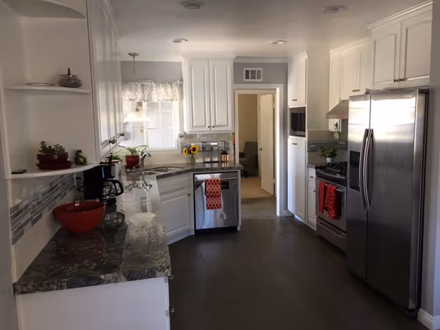 A modern kitchen with white cabinets, granite countertops, and stainless steel appliances including a refrigerator, stove, and dishwasher. There are red kitchen towels hanging on the dishwasher and stove. A window with a valance lets in natural light above the sink. The floor is dark wood, and there are small plants and kitchen items on the counters.