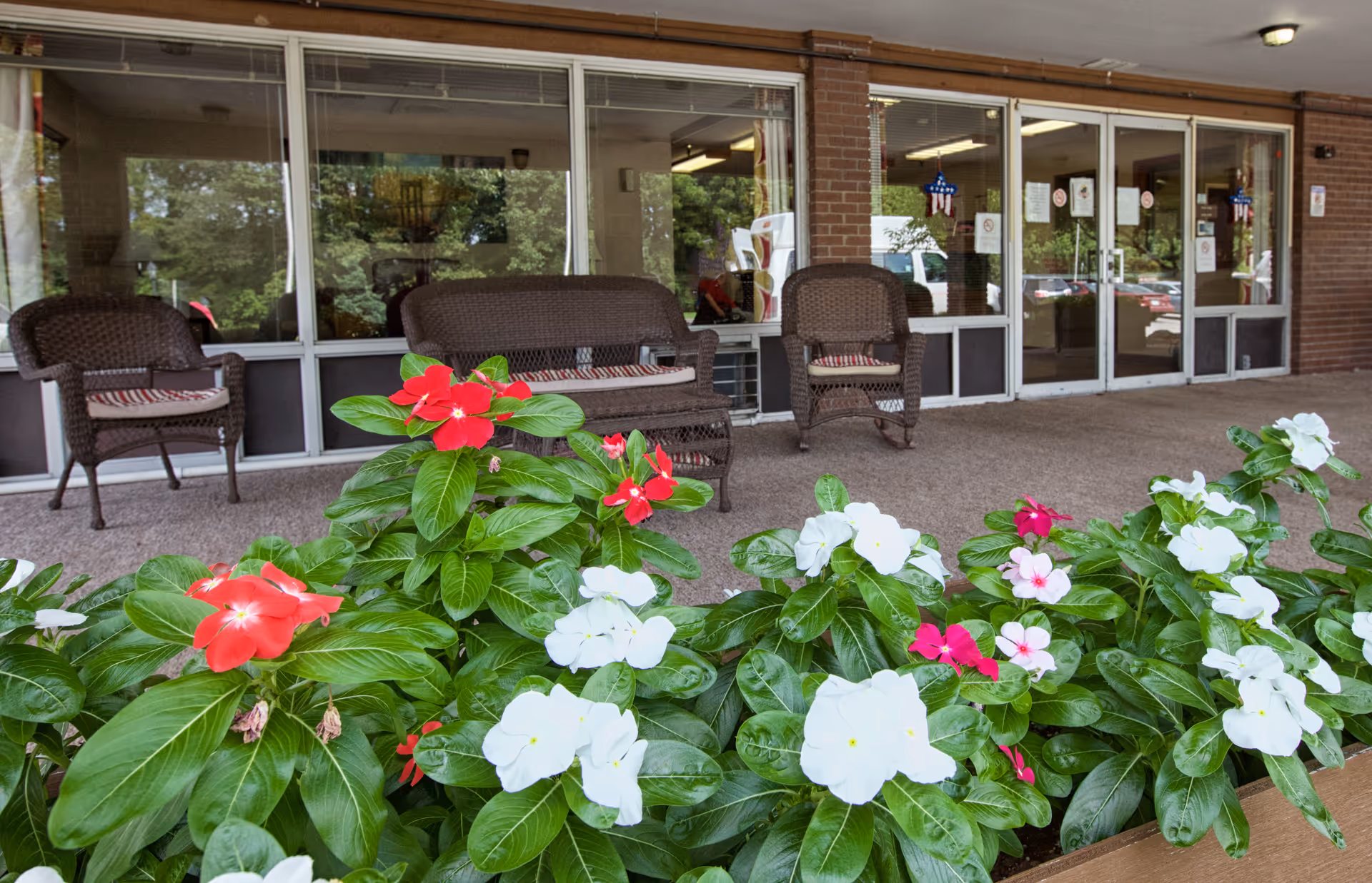 Outdoor seating area with brown wicker chairs and a loveseat with cushions, situated on a covered patio in front of large windows and glass doors. In the foreground, there is a planter box filled with vibrant red, white, and pink flowers with green leaves.