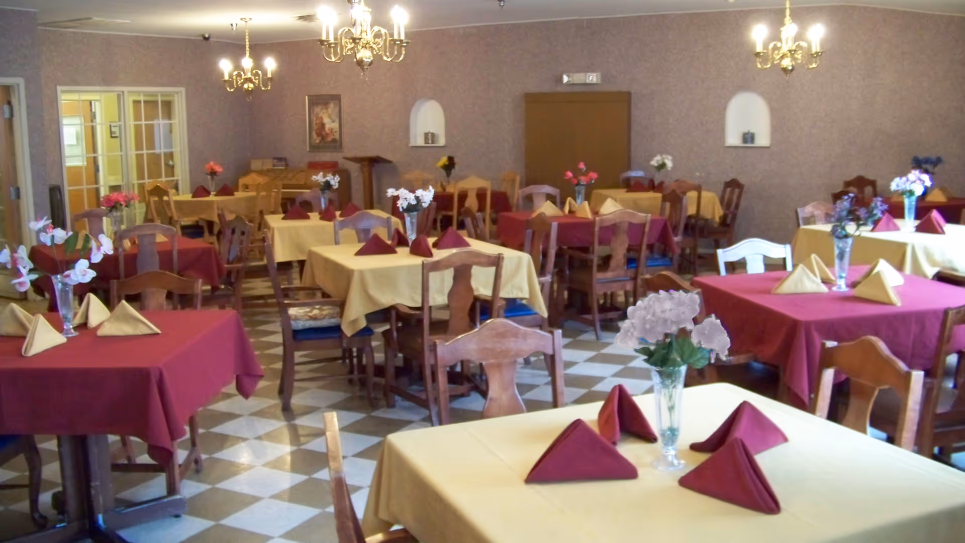 Dining room with multiple tables covered in red and yellow tablecloths, folded napkins, and floral centerpieces.