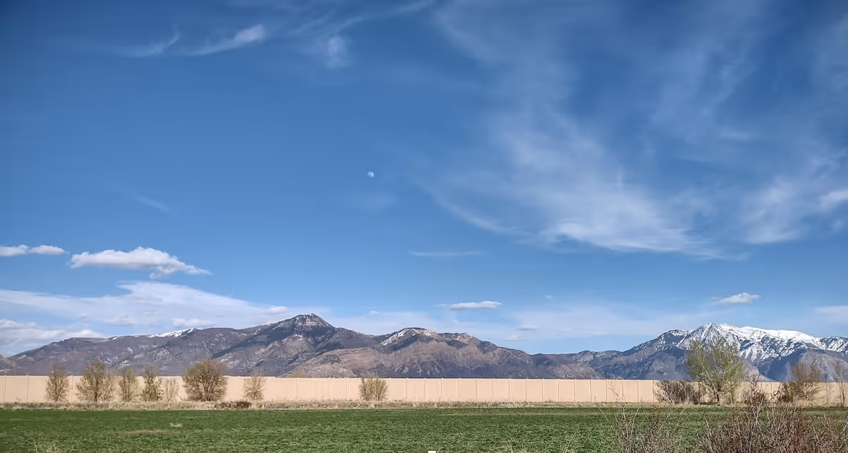 A scenic outdoor view featuring a green grassy field in the foreground, a row of small trees and shrubs, a beige wall or fence, and a range of mountains in the background under a blue sky with scattered clouds and a visible moon.