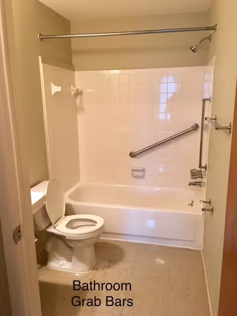 A bathroom with a white toilet, a bathtub with white tiled walls, and multiple grab bars installed on the walls around the bathtub. There is a shower curtain rod above the bathtub and a beige floor.