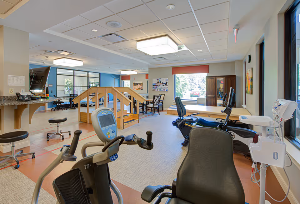 Interior view of a rehabilitation facility room with exercise equipment including stationary bikes, a set of wooden stairs for physical therapy, chairs, tables, and large windows letting in natural light.