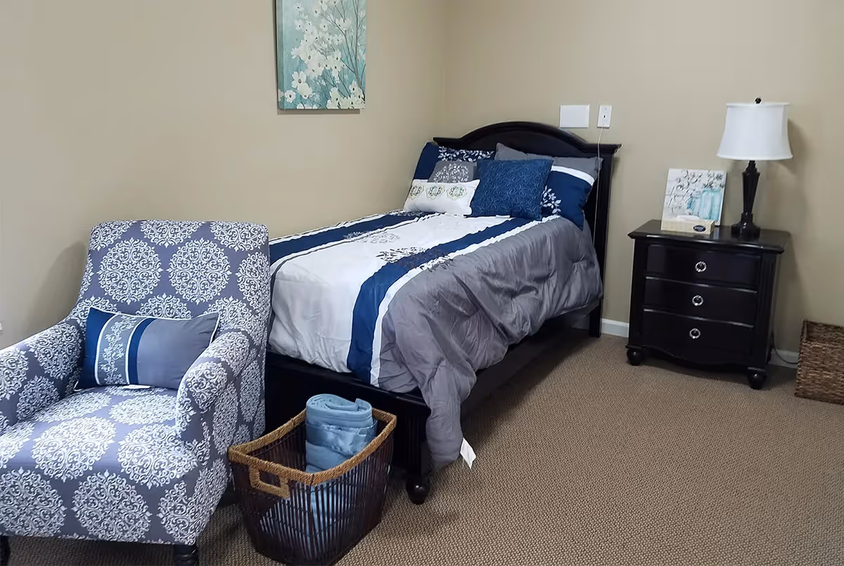 A tidy bedroom with a single bed dressed in blue and gray linens, a patterned armchair, nightstand with a lamp, and a basket on a carpeted floor.