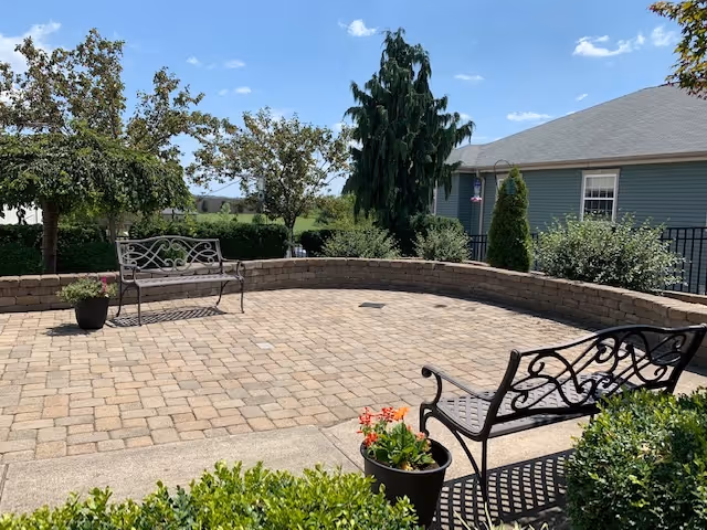 Outdoor patio area with two decorative metal benches, potted plants, and a low stone wall surrounding the paved space. Trees and shrubs are visible in the background along with a blue building under a clear sky.