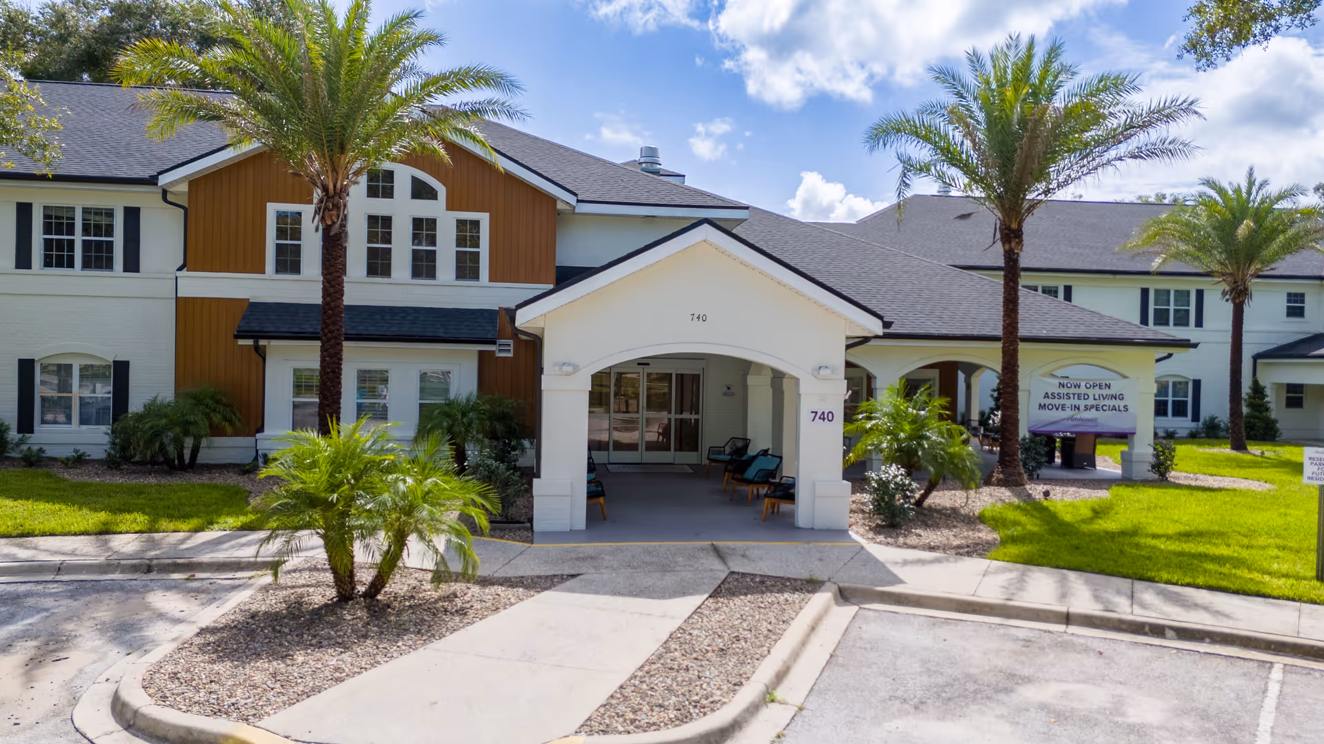 Front exterior view of Ambiance at Maitland senior living facility with a covered entrance, palm trees, green lawn, and a sign advertising assisted living move-in specials.