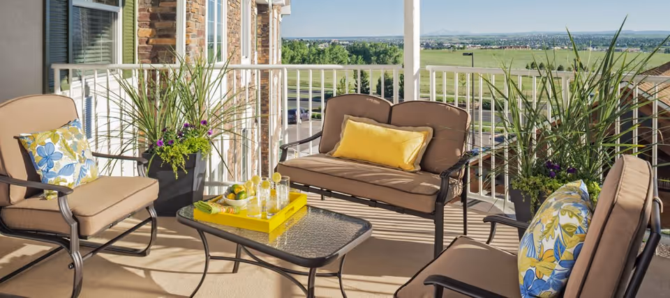 Outdoor patio area with cushioned seating including two armchairs and a loveseat, decorated with colorful floral and yellow pillows. A glass-top coffee table holds a yellow tray with glasses and lemons. Potted plants are placed near the white railing, overlooking a green landscape and distant hills under a clear sky.