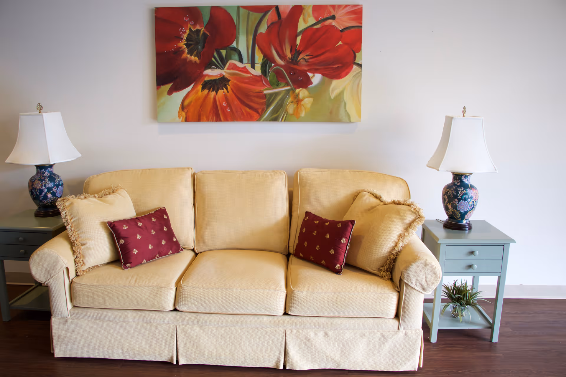 A beige sofa with two beige and two red decorative pillows is positioned against a white wall. On either side of the sofa are small green side tables, each with a blue floral lamp with white lampshades. Above the sofa hangs a colorful painting of red and orange flowers.