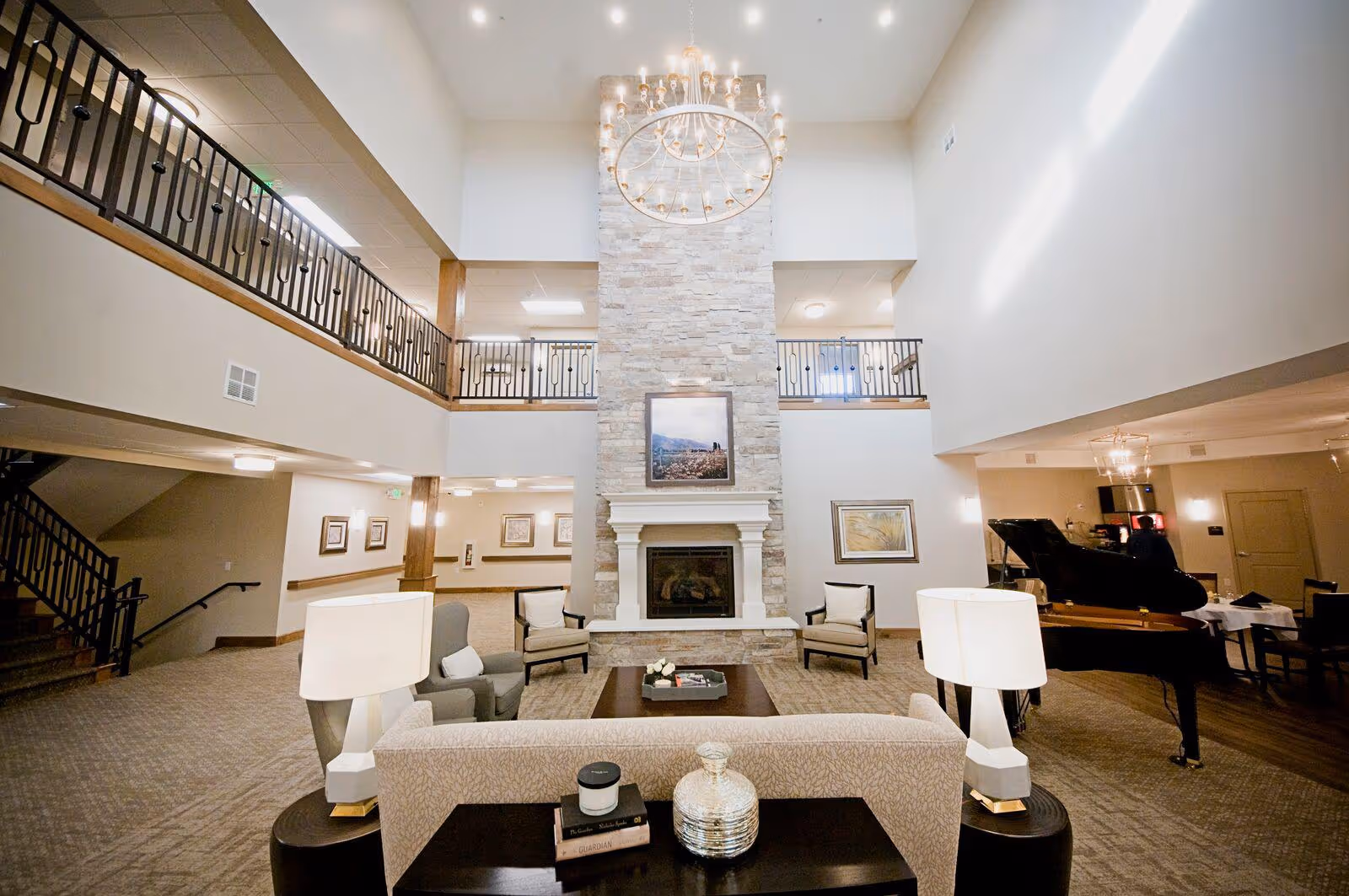 Spacious and well-lit living room area in Blacksmith Fork Assisted Living featuring a tall stone fireplace with a framed picture above it, a large chandelier hanging from the high ceiling, comfortable seating including armchairs and a sofa, a grand piano to the right, and a staircase leading to an upper balcony.