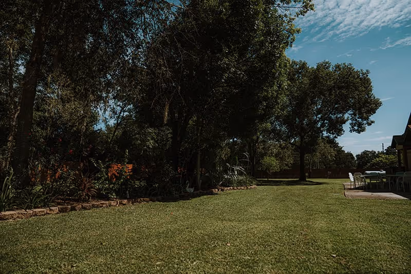 A spacious, well-maintained outdoor garden area with green grass, large trees providing shade, and a patio area with tables and chairs under a partly cloudy sky.