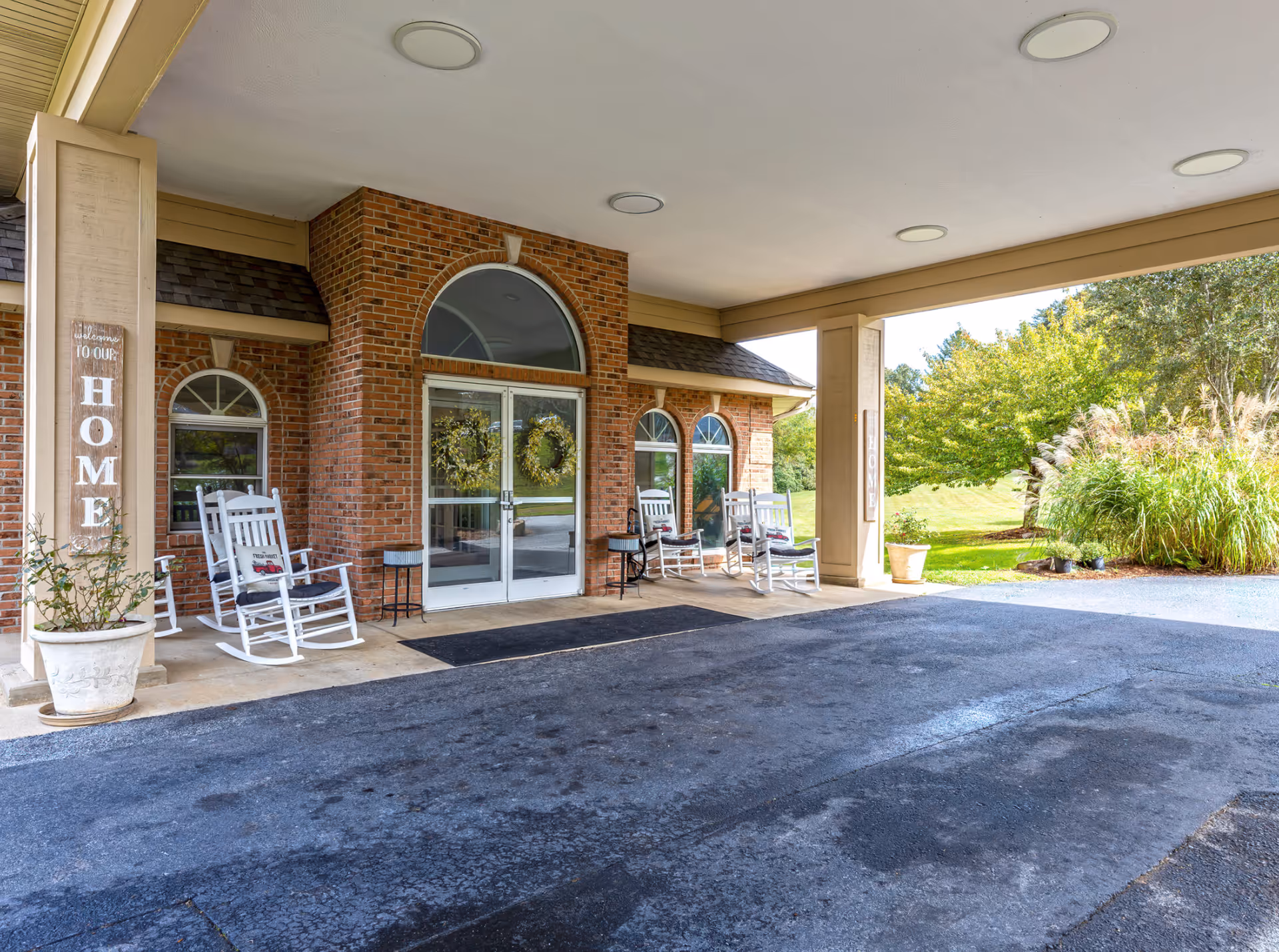 Covered entrance area of a brick building with glass double doors decorated with wreaths. There are white rocking chairs with cushions on either side of the entrance and potted plants. The pillars have vertical signs that say 'Welcome to our HOME'. Green trees and bushes are visible in the background.
