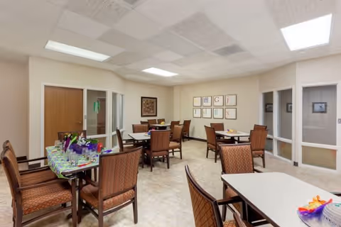 Communal dining room with several tables and upholstered chairs arranged under fluorescent ceiling lights.