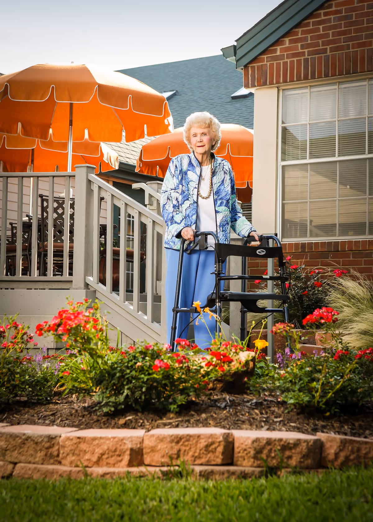 An elderly woman with white hair using a walker stands outside near a garden with colorful flowers. Behind her is a building with brick walls and large windows, and several orange patio umbrellas are visible in the background.