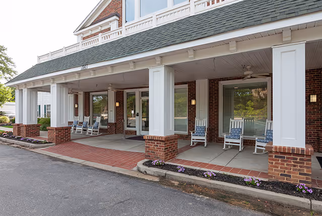 Front entrance of a brick assisted living community building with a covered porch supported by white columns. Several white rocking chairs with blue cushions are arranged on the porch. There are small flower beds with purple flowers along the edge of the porch.