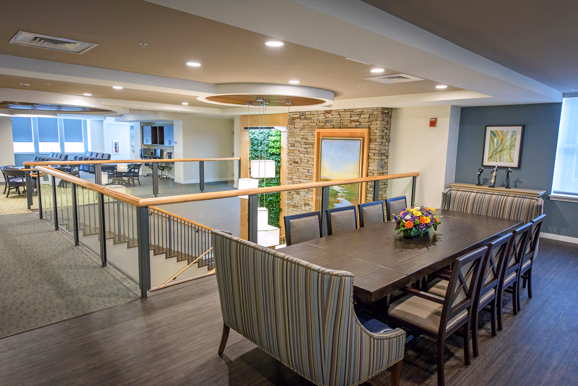 Bright, modern communal dining area with a long wooden table, upholstered chairs and bench seating, a floral centerpiece, and a railing overlooking a lower level.