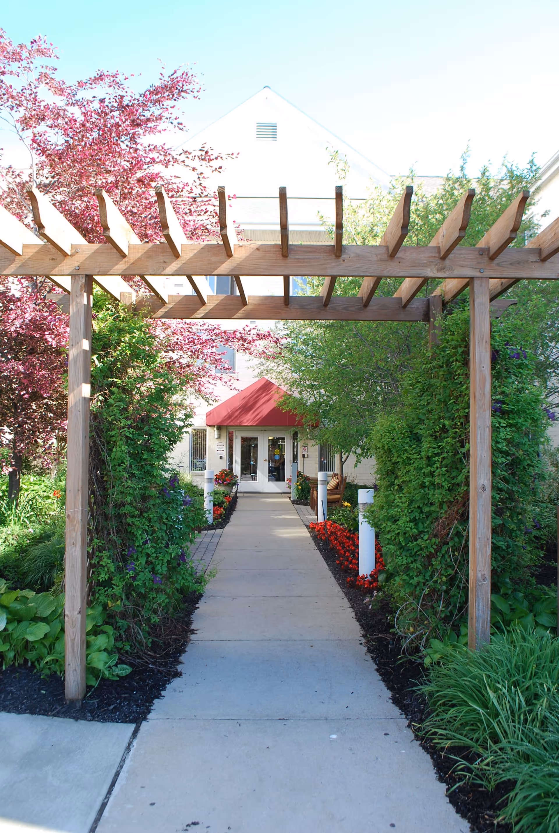 A walkway under a wooden pergola leads through landscaped greenery and flowers to a building entrance with a red awning.