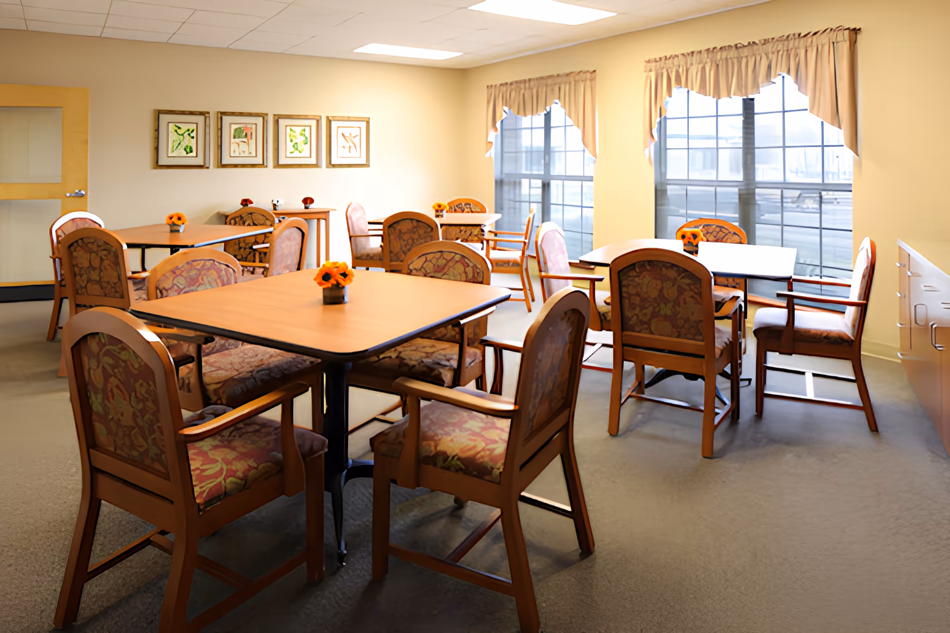 Bright communal dining room with wooden tables and upholstered chairs, small flower centerpieces, and large windows.