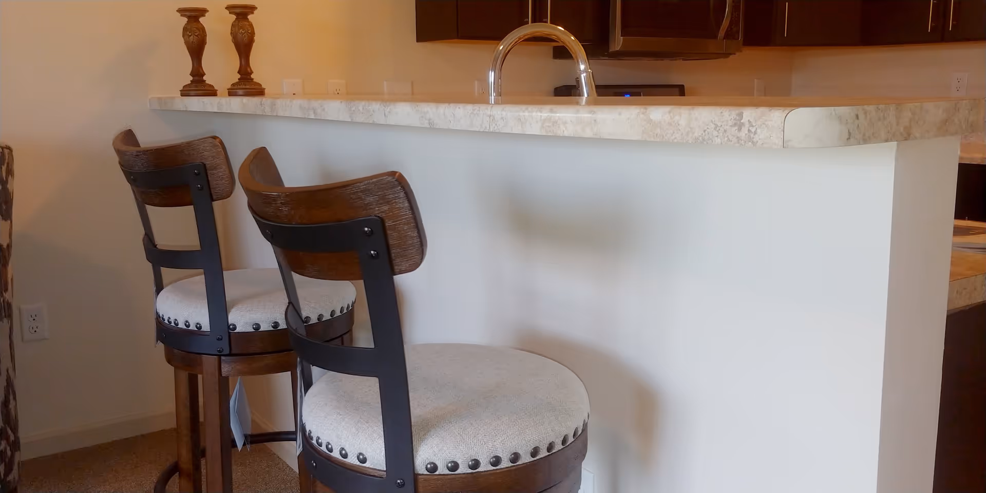 Two wooden bar stools with cushioned seats positioned at a kitchen counter with a marble-like surface. The counter has a modern faucet, and there are two decorative candle holders on the counter. The background shows part of a kitchen with dark cabinets and a microwave.