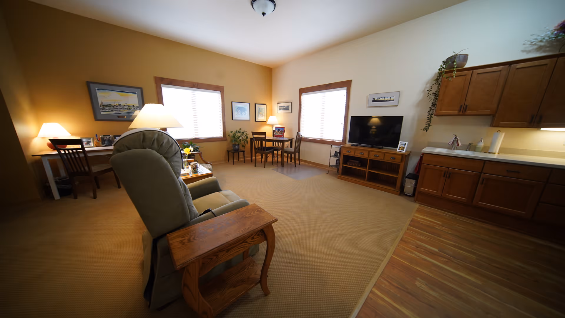 A cozy living room area with a recliner chair and wooden side table in the foreground. The room has beige carpet and wood flooring near a kitchenette with wooden cabinets and a countertop. There is a TV on a wooden stand, two windows with blinds, a small dining table with chairs, and a desk with a chair and lamp. The walls are painted in warm tones with framed artwork and some plants.