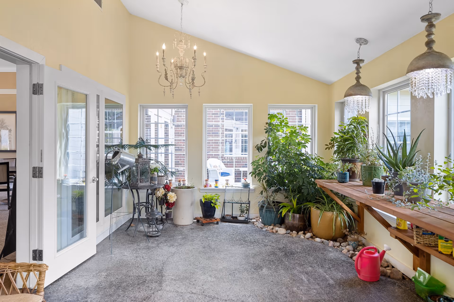 A bright indoor garden room with large windows letting in natural light. The room features various potted plants on the floor and on a wooden shelf along the right wall. There are two hanging pendant lights with crystal decorations and a chandelier hanging from the ceiling. French doors on the left side open into another room.
