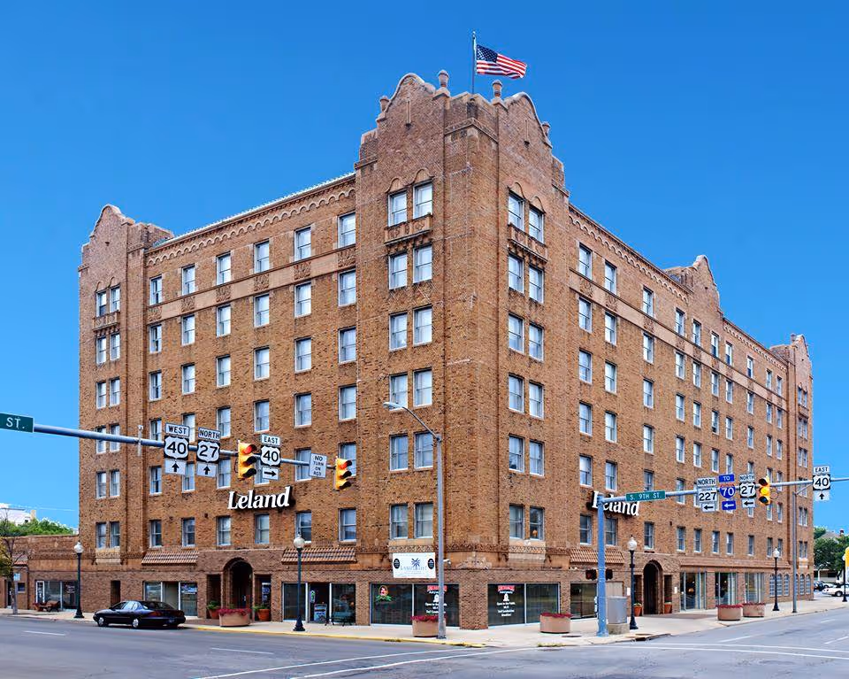 Exterior view of a large, multi-story brick building with an American flag on top, located at a street intersection with traffic lights and street signs. The building has multiple windows and the name 'Leland' displayed on the facade.