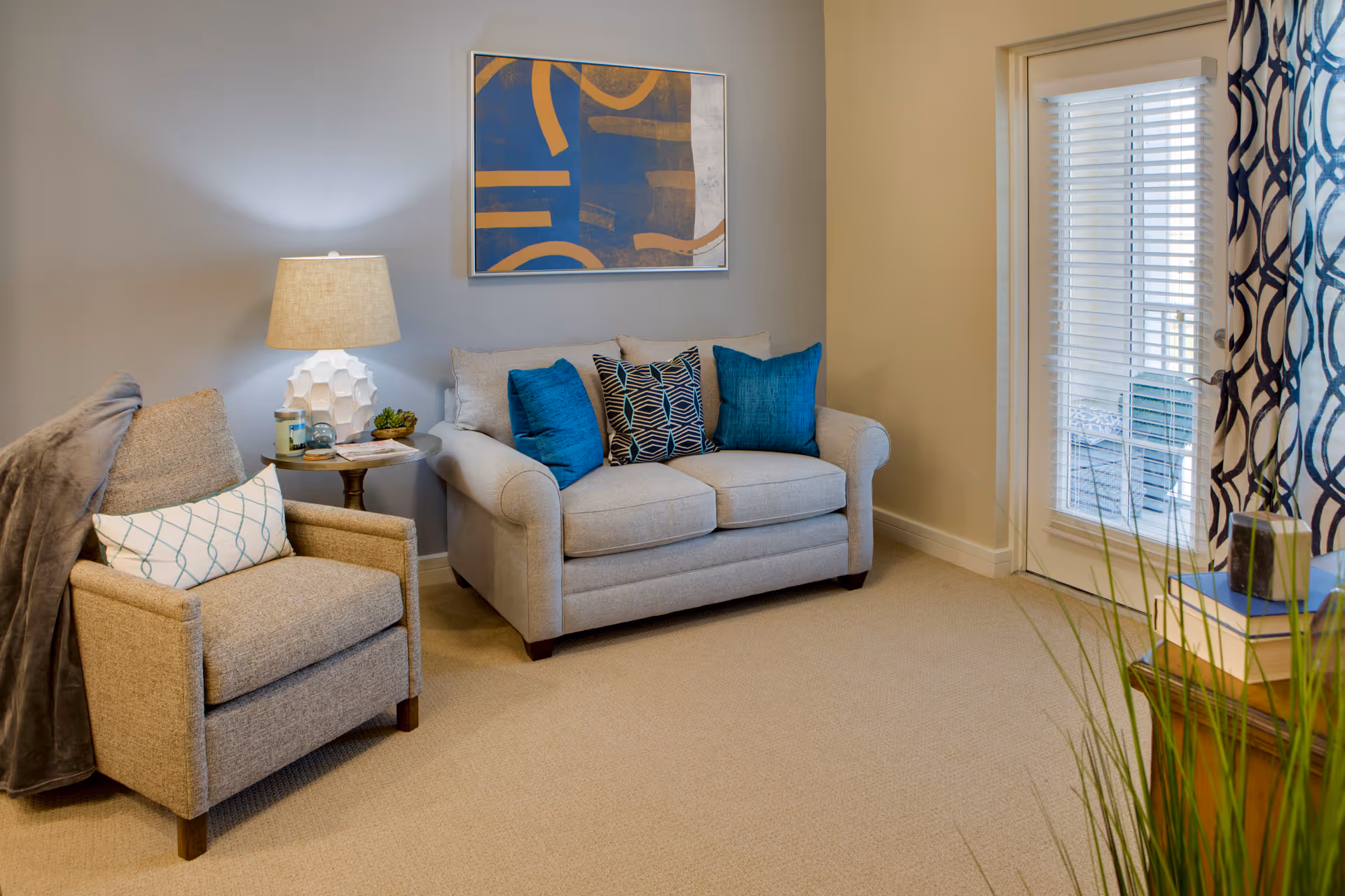 A cozy living room with a beige loveseat adorned with three decorative pillows in blue and patterned designs, a beige armchair with a white pillow and a gray throw draped over the side, a round side table with a white textured lamp, candle, and small plant, a framed abstract artwork on the wall, and a glass door with white blinds and patterned curtains leading outside.