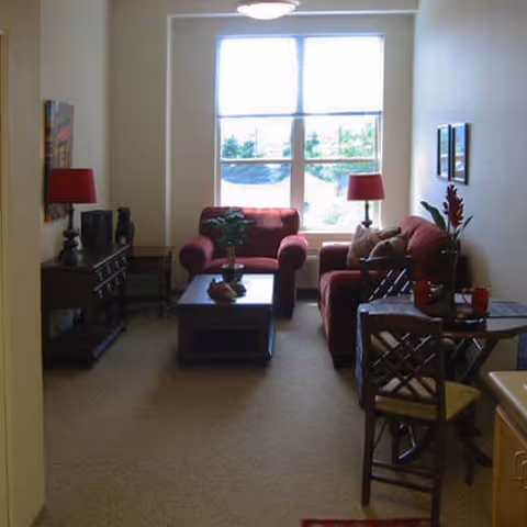 A cozy living room with two red upholstered armchairs and a matching sofa arranged around a dark wooden coffee table. There are two red lamps on side tables, a small dining table with two chairs, and a large window letting in natural light. The walls are light-colored and decorated with framed pictures.