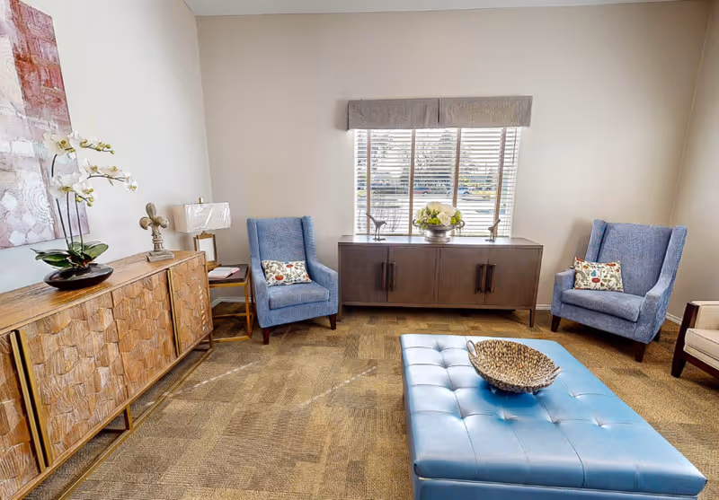 A cozy living room area with two blue upholstered armchairs each with a decorative pillow, a wooden sideboard with a lamp and a plant, a large window with blinds, and a blue tufted ottoman with a decorative bowl on top.
