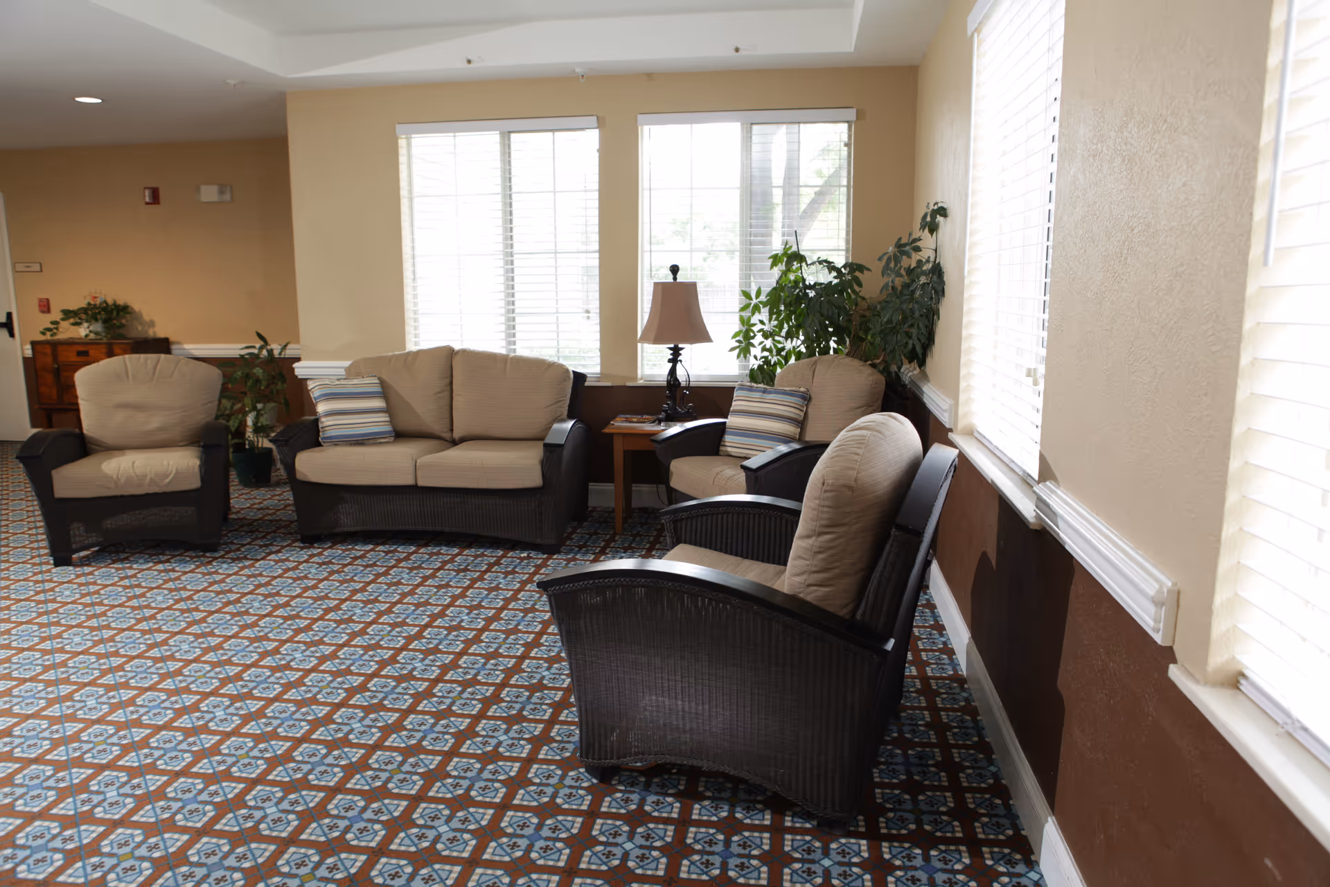 A cozy sitting area in a senior living facility with four cushioned wicker chairs arranged around a small wooden side table with a lamp. The room has large windows with white blinds, beige walls, and a patterned carpet floor. There are also some green plants near the windows.