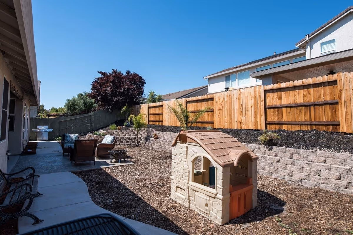 Outdoor patio area with a small plastic playhouse, seating area with chairs and cushions, a fire pit, and a barbecue grill. The space is enclosed by a wooden fence and has a clear blue sky overhead.