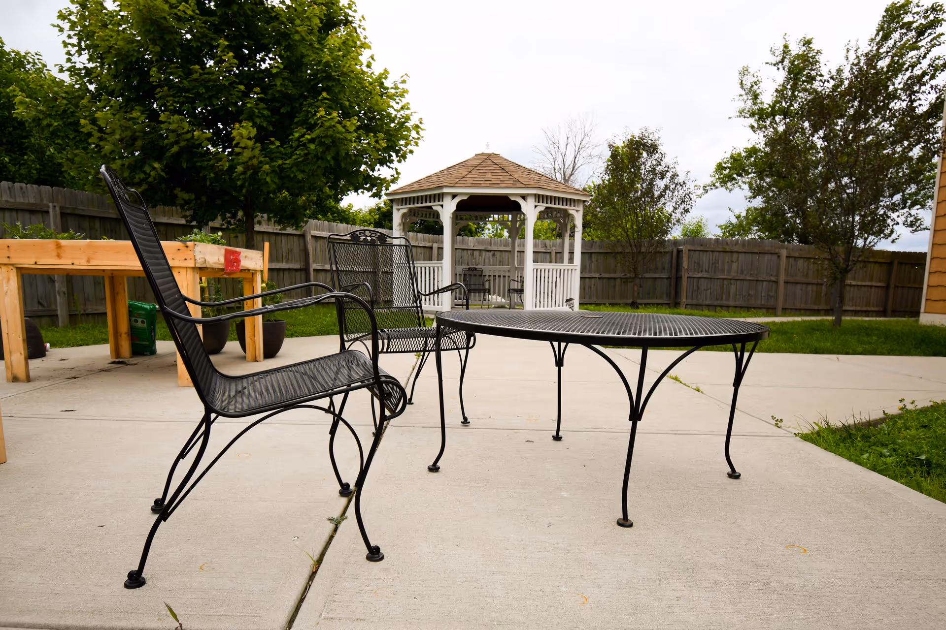 Outdoor patio area with black metal chairs and a matching round table on a concrete surface. In the background, there is a white gazebo surrounded by a wooden fence and green trees.