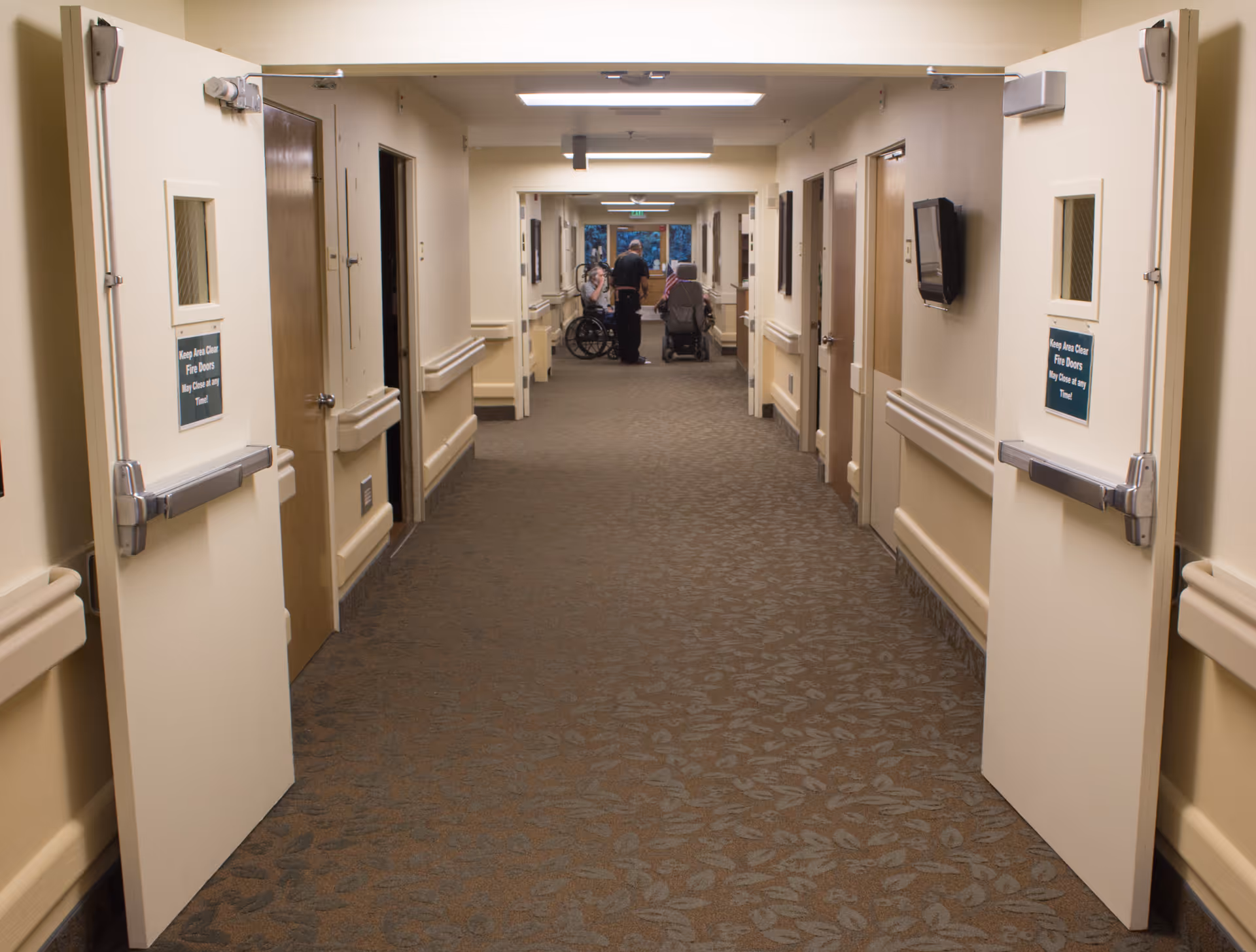 A long hallway in a senior living facility with beige walls and patterned carpet. Double doors are open at the near end of the hallway. Several closed doors line both sides of the hallway. At the far end, three people are visible, two seated in wheelchairs and one standing, engaged in conversation.