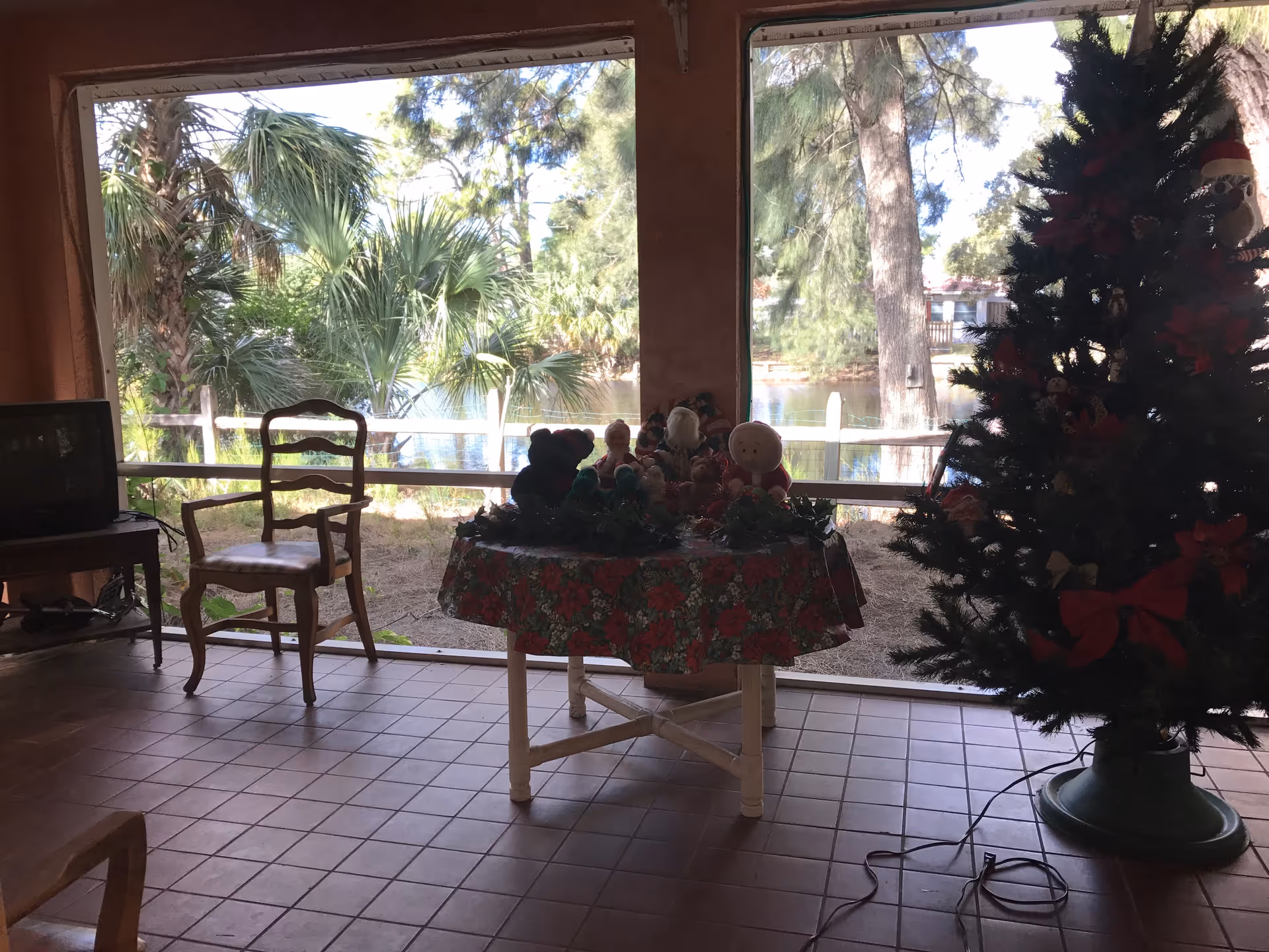 Interior view of a room with large windows showing an outdoor scene with trees and a body of water. Inside, there is a wooden chair, a small table covered with a festive tablecloth and holiday decorations, and a decorated Christmas tree. A small television is on a stand to the left.