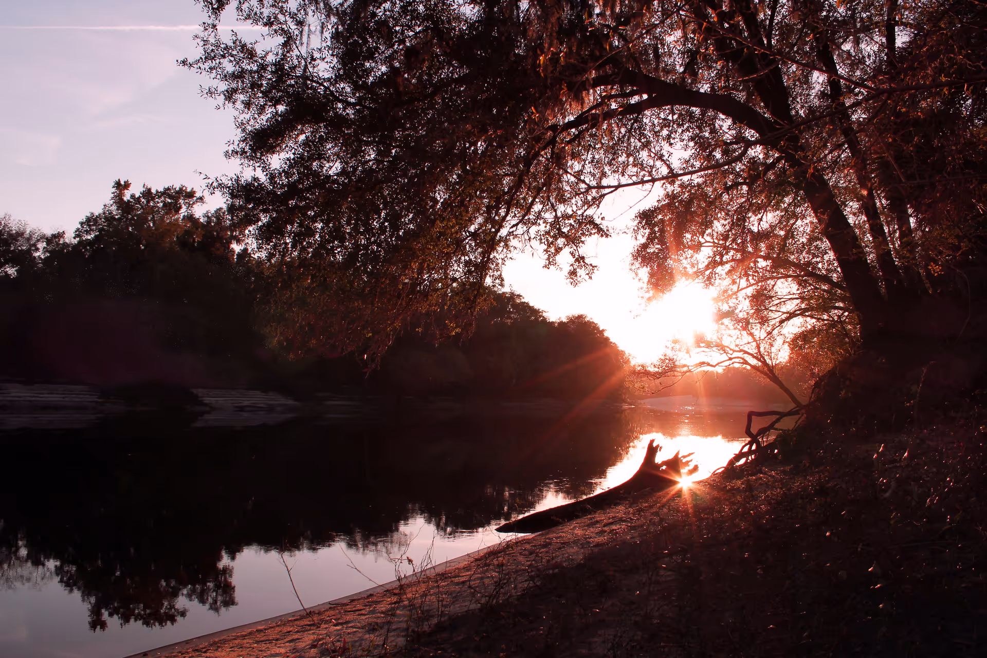 Sunset over a calm river with trees on both sides and a sandy riverbank in the foreground, with sunlight filtering through the branches.