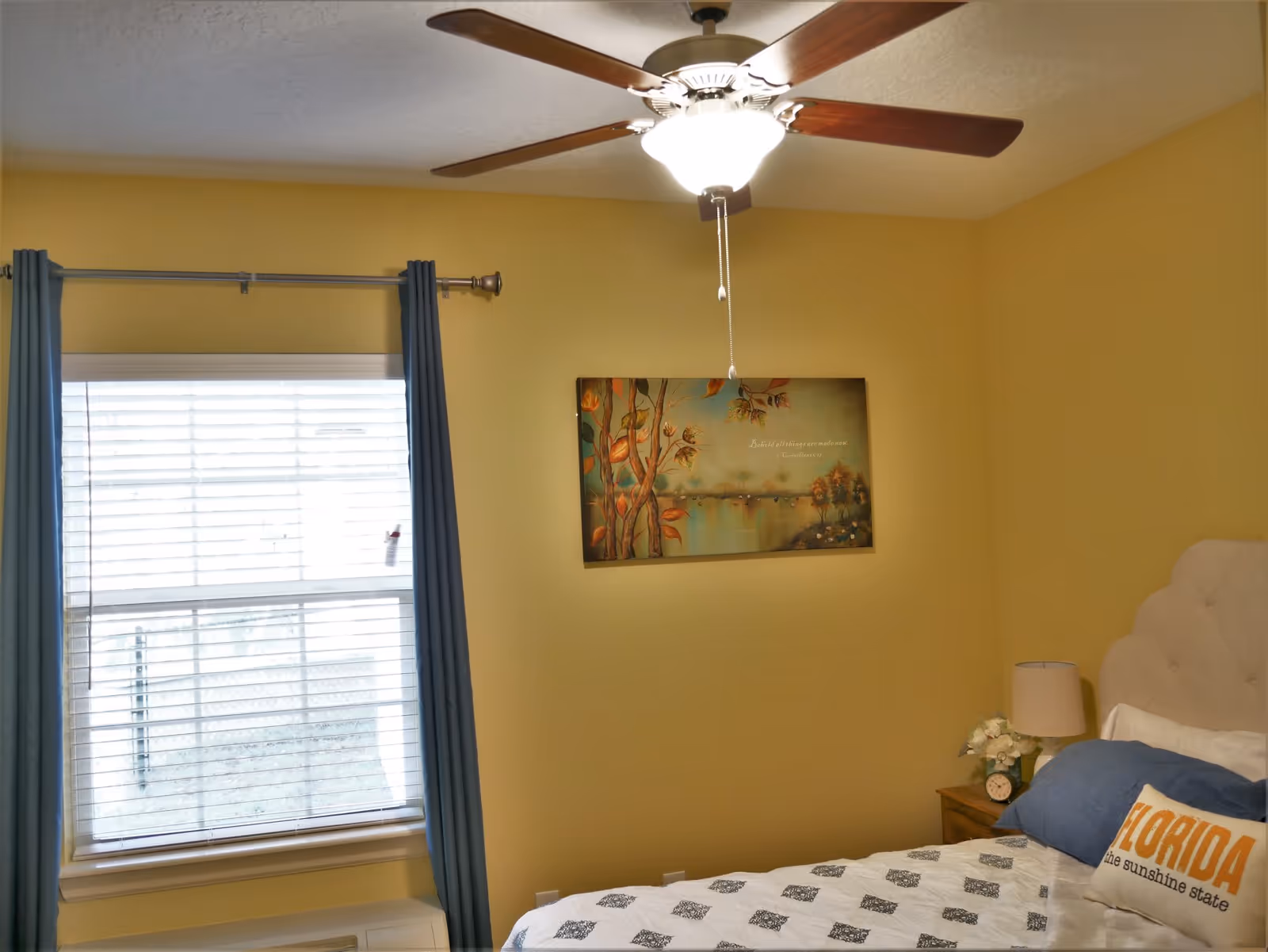 A cozy bedroom with yellow walls, a window with blue curtains, a ceiling fan with lights, a bed with white and blue bedding, and a pillow that says 'FLORIDA the sunshine state'. There is a nightstand with a lamp, a clock, and a small flower arrangement. A painting with autumn leaves and a lake scene hangs on the wall.