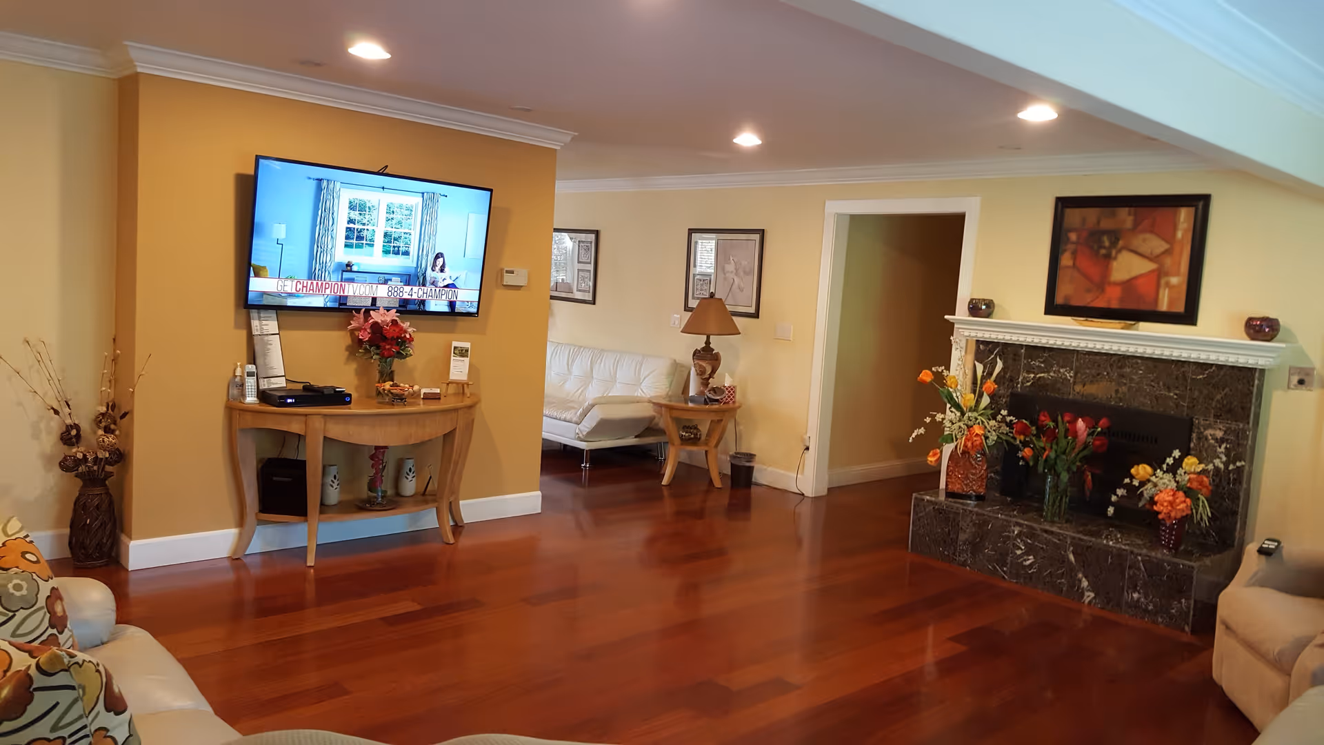A cozy living room with polished wooden floors, a mounted flat-screen TV displaying a commercial, a wooden console table with decorative items and flowers, a white leather sofa, a side table with a lamp, and a marble fireplace adorned with multiple flower arrangements and a framed abstract painting above it.