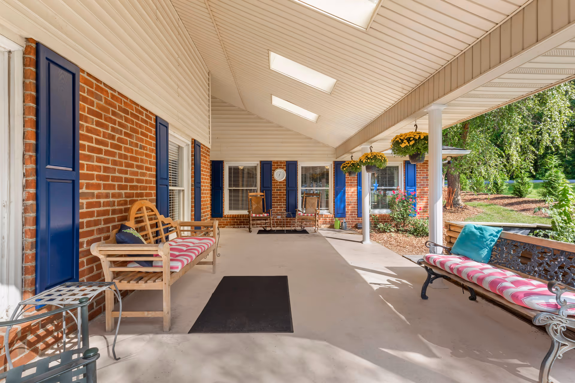 Covered outdoor patio area with brick walls and blue shutters. There are wooden and metal benches with striped cushions, hanging flower pots with yellow flowers, and a view of a garden with trees and shrubs.